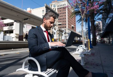 Takım elbiseli olgun bir işadamı açık havada bir bankta oturuyor. İnternette, dizüstü bilgisayarda çalışıyor.