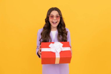 Güneş gözlüklü ve takım elbiseli son moda çocuk hediye kutusunu sarı arka planda tutuyor.