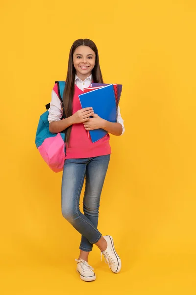 Happy teen school girl with backpack and copybook on yellow background, education — Zdjęcie stockowe