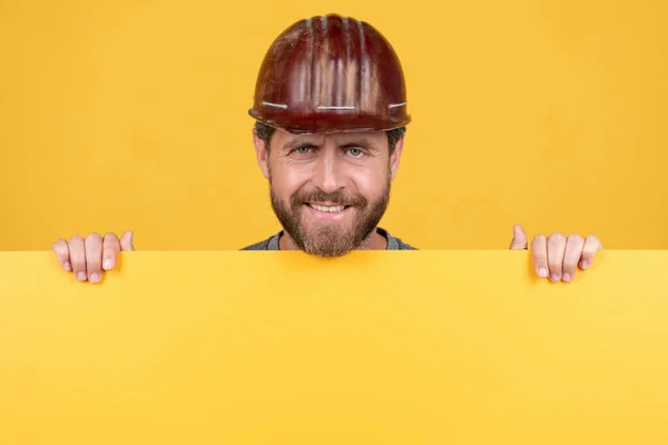 Happy mature man constructor in helmet behind yellow paper for copy space, happy labor day — Stock Photo, Image