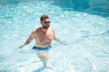 Yaz tatilinde havuzda yüzen güneş gözlüklü mutlu sakallı adam.