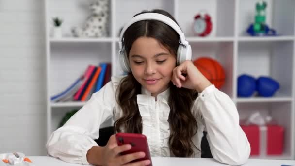 Colegiala alegre en auriculares comprobar el correo electrónico y el mensaje de lectura en el teléfono, chat de la escuela. — Vídeos de Stock