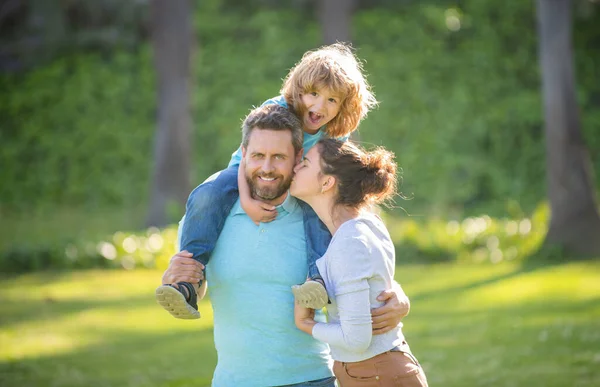 Mother kiss father while boy child riding piggy back on fathers shoulders summer outdoors, family — Stock Photo, Image