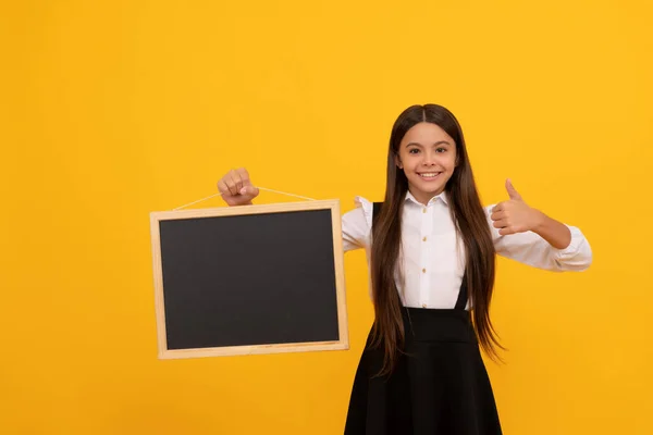 Menina adolescente alegre em uniforme segurar escola quadro negro para espaço de cópia mostrando polegar para cima, apresentando — Fotografia de Stock