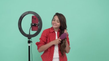 Gülümseyen çocuk güzellik blogcusu saç fırçasını tanıtıyor ve uzun dalgalı saçlarını tarıyor, terfi ettiriyor.