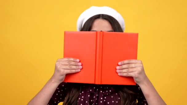 Умная школьница в очках прячется за тетрадью твердой обложки для чтения и учебы, литературы — стоковое видео