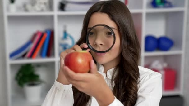 Uśmiechnięty dzieciak w szkolnym mundurze spoglądający przez szkło powiększające na jabłko, odkrycie — Wideo stockowe