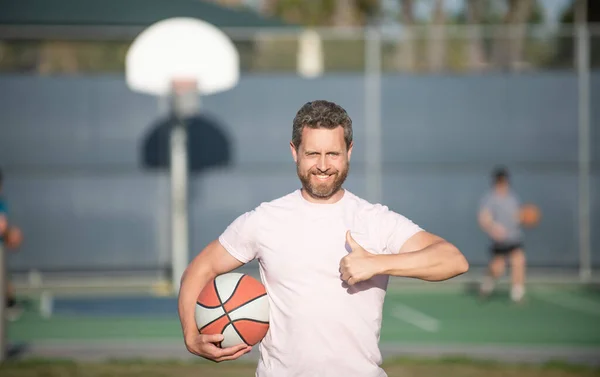 Alegre hombre muscular atleta baloncesto jugador celebrar pelota al aire libre mostrando pulgar hacia arriba, el éxito deportivo. — Foto de Stock