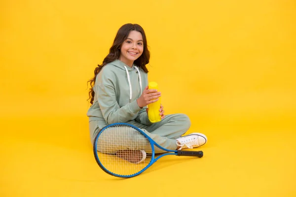 Bambino felice tenere racchetta da tennis e bere acqua dalla bottiglia su sfondo giallo, idratazione. — Foto Stock