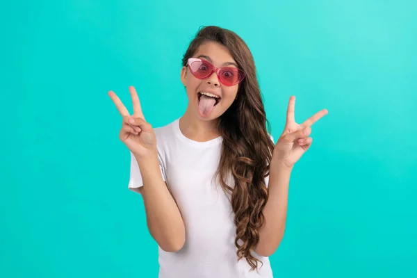 Srandovní teen dívka dlouhé kudrnaté vlasy v slunečních brýlích ležérní styl na modrém pozadí mír gesto, zábava — Stock fotografie