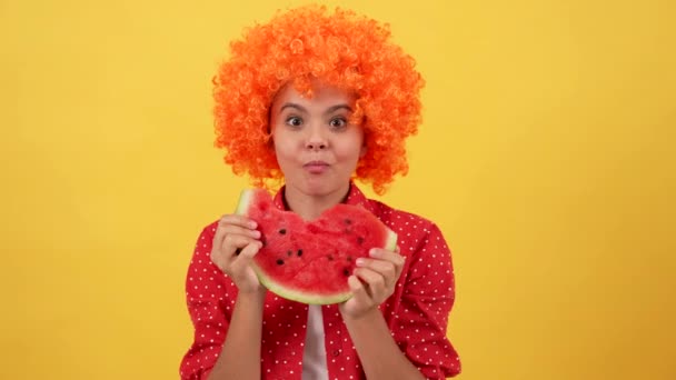 Happy hungry teen girl in orange hair wig showing tongue and biting water melon slice, summer — Stock Video