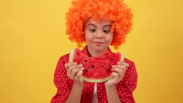 Κατάπληκτος πεινασμένο παιδί σε φανταχτερό πορτοκαλί περούκα τρώνε καρπούζι φέτα σε κίτρινο φόντο, πείνα — Αρχείο Βίντεο