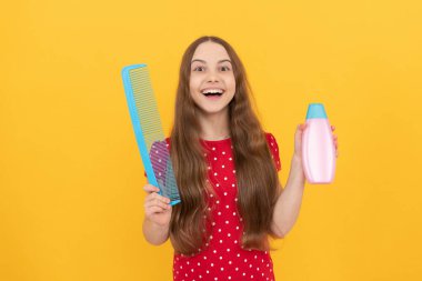 Şaşırmış çocuk, sarı arka planda uzun saçlar için şampuan ve saç fırçası sunuyor.