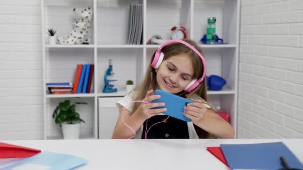 Colegiala feliz en los auriculares viendo vídeo musical en el teléfono celular en el aula, estudiar en línea — Vídeos de Stock