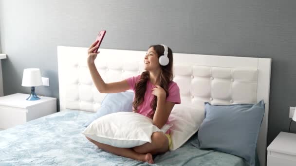 Glückliches Kind mit Kopfhörer macht Video-Selfie auf Handy entspannt im Bett, moderne Bildung — Stockvideo