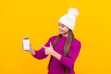 Doğal hap ürünleri olan genç bir kız. Vitamin ürünü sunuyorum. Çocuklar için multivitamin tableti. Sağlıklı bir yaşam tarzı. Çocukluk sağlığı. Kışın şapka takan çocuk hap kavanozunu işaret ediyor. yiyecek takviyesi.