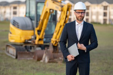 İnşaat işyeri sahibi. İnşaat alanında takım elbiseli ve şapkalı bir adam. İnşaat mühendisi kazıcının yanındaki evin önünde. Mimar, yönetici, yatırımcı, inşaat yöneticisi