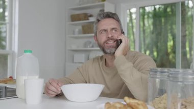 Çevrimiçi işleri yönetirken kahvaltı yapan İspanyol adam. Günaydın merkez ofis. Modern mutfakta sabah kahvaltısı. Mısır gevreği yiyen adam telefonla konuşuyor. Adam sabah kahvaltısını mutfakta yiyor..