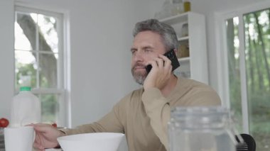 Adam sabah kahvaltısını mutfakta yiyor. Çevrimiçi işleri yönetirken kahvaltı yapan adam. Günaydın merkez ofis. Modern mutfakta sabah kahvaltısı. Mısır gevreğini sütle yiyen ve telefonla konuşan bir adam..