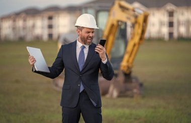 Takım elbiseli ve kasklı bir Buider. İnşaat yatırımcısı. İnşaat alanının önündeki yatırımcı. Başarılı bir yatırımcı. İnşaatta takım elbiseli yakışıklı ve kasklı bir adam.