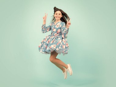 Uzun boylu, neşeli genç kız 1213 yaşında, mavi arka plan stüdyosunda tek başına atlayan. Komik atlayış. Yazlık hareket elbisesi içindeki çocuk