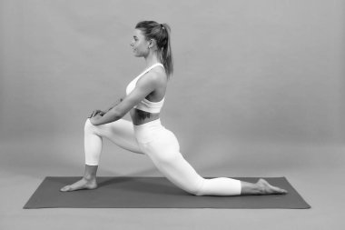 Sportif kız, yoga minderi üzerinde egzersiz yaparken, bacaklarını yukarı kaldırıyor, arka planda gri bir alan var.