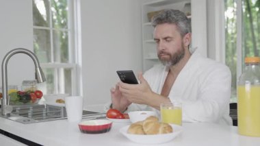 Sabahları sağlıklı bir kahvaltı. Mutfakta telefonla kahvaltı eden olgun bir adam, sağlıklı bir yaşam tarzı. İspanyol adam kahvaltı yapıyor. Sabah yemeği. İnsan yemek yer. Sabah kahvaltısı.