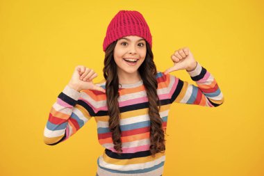 Beautiful teen girl in a winter hat and a warm sweater. A child on a yellow isolated background. Happy teenager, positive and smiling emotions of teen girl