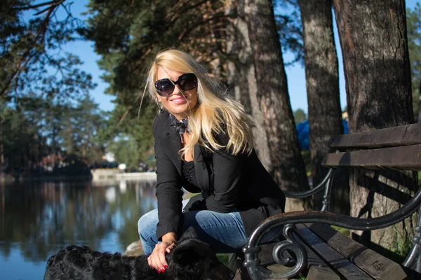 Young pretty girl with blonde hair and black glasses, smiling in park with a dog. Beautiful blond hair, photographed on a bench in park. — Stok fotoğraf