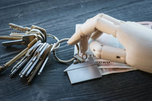 Mão de madeira artificial segurando as chaves e dinheiro, em uma mesa de madeira — Fotografia de Stock