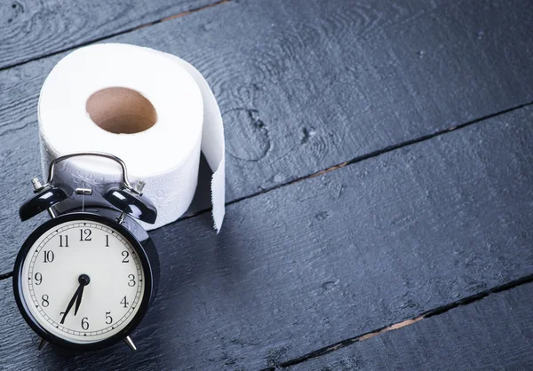 Alarm clock with toilet paper on a black wooden table — Stock Photo, Image