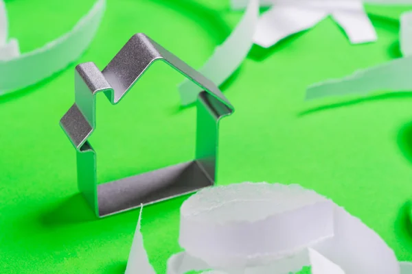 Metal house standing on a green surface with teared paper pieces — Stock Photo, Image