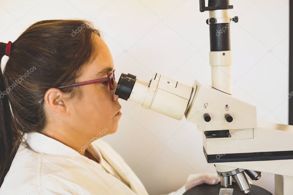 scientist with her white apron sitting on a chair with her red glasses in her laboratory observing a sample through a microscope