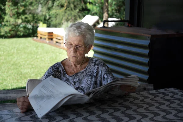 Gray-haired and short-haired old woman with glasses. The woman is alone. The elderly lady is sitting reading a newspaper in the garden of her house. The ancient is wearing a black and white shirt. Lay the journal on a large table. The day is sunny so