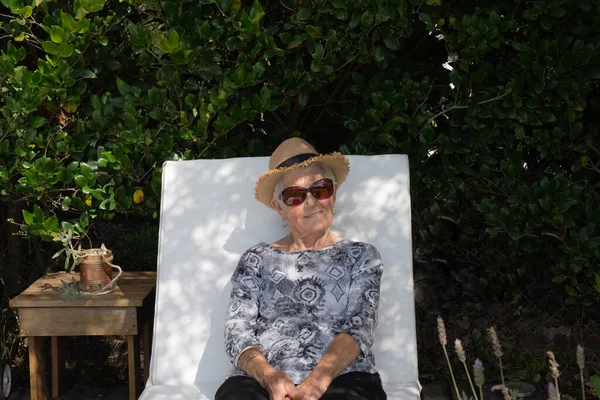 身材矮小 头发灰白 头戴太阳镜 头戴帽子的成年妇女 那个古代人正坐在她家花园的一张躺椅上 那个女人孤身一人 很快乐 穿着一件黑白T恤 天气很好 所以有许多自然光 — 图库照片