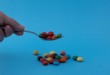 Kaşık tutan bir elin yakın çekimi. Kaşığın üzerinde renkli şekerler var. Mavi arka planda daha renkli şekerlemeler de görülebilir.