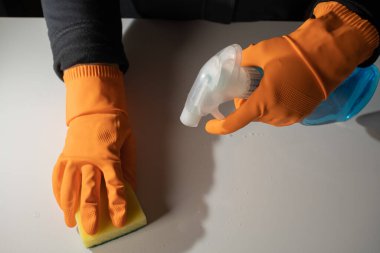 Turuncu eldivenli eller, süngerle masayı temizliyor ve temizlik malzemesi ile kapatılıyor..