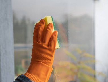 Elleri turuncu temizleme eldiveniyle pencerenin camını temizliyor. Arka planda çalıştığınız yerin bahçesini görebilirsiniz..