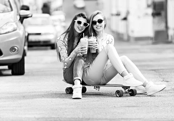 Όμορφα νεαρά κορίτσια hipster φίλες θέτουν με skateboard με το κάθισμα στο skate, Οδός μόδα τρόπο ζωής στο γυαλιά ηλίου. Κρατήσει κοκτέιλ και χαμογελαστή — Φωτογραφία Αρχείου