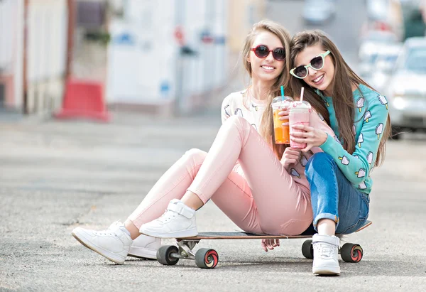 Vackra unga flickor hipster flickvänner poserar med en skateboard säte på skate, street mode livsstil i solglasögon. Hålla cocktail och leende — Stockfoto