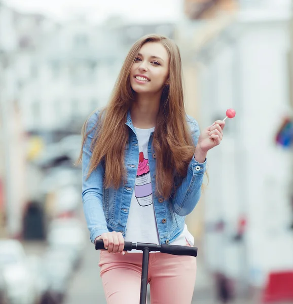 Hipster fashion girl urban style . Candy in a hand on scooter in the city — Stock Photo, Image