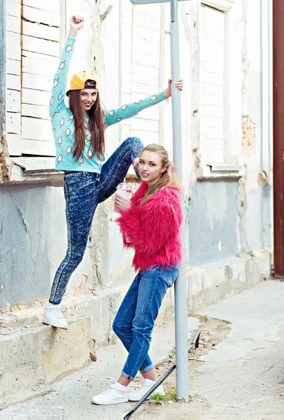 Color fashion portrait of beautiful glam street style girlfriends standing near grange wall. keep cocktail in a hand — Stock Photo, Image