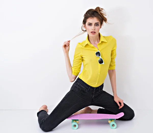 Moda hipster ragazza in camicia gialla in occhiali da sole con skateboard flirtare contro il muro bianco . — Foto Stock