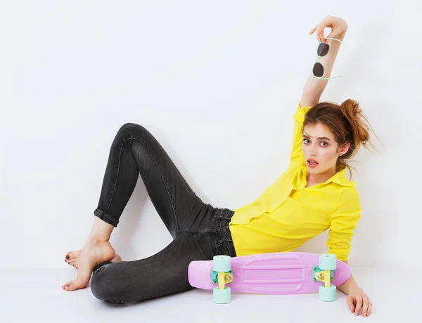 Moda chica hipster en camisa amarilla en gafas de sol con monopatín coqueteando contra la pared blanca . — Foto de Stock