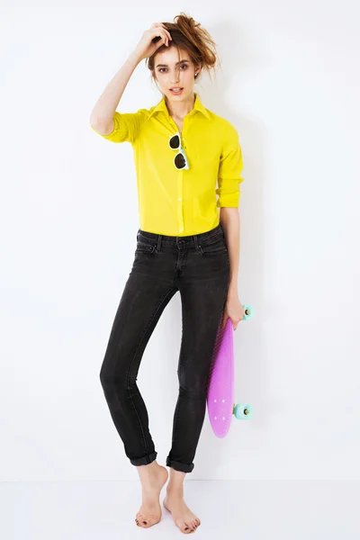 Mode-Hipster-Mädchen im gelben Hemd mit Sonnenbrille und Skateboard flirtet gegen die weiße Wand. — Stockfoto