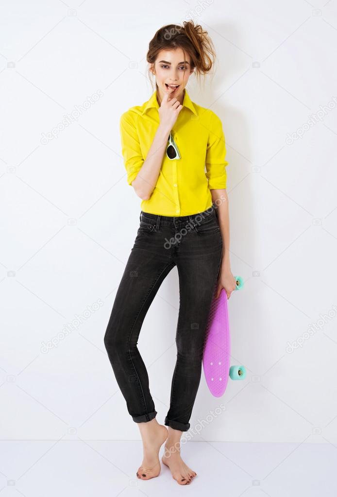 Fashion hipster girl in yellow shirt in sunglasses with skateboard flirting against the white wall.