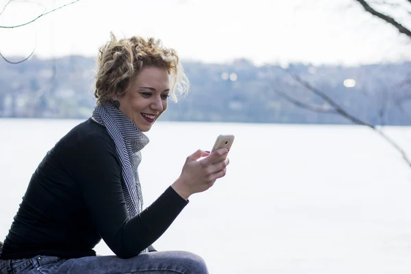 Heureuse jeune femme blonde bouclée assise au bord de la rivière et tapant sur — Photo