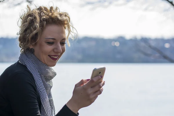 Heureuse jeune femme blonde bouclée assise au bord de la rivière et tapant sur — Photo