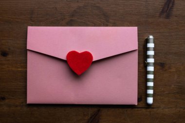 Üzerinde kırmızı bir kalp bulunan bir düğün davetiyesi olan pembe renkli bir zarf koyu ahşap bir masanın üzerindeki küçük altın ve beyaz bir kalemin yanında, Toronto, Ontario, Kanada.