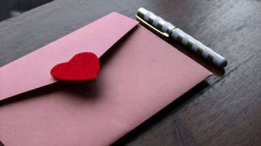 Üzerinde kırmızı bir kalp bulunan bir düğün davetiyesi olan pembe renkli bir zarf koyu ahşap bir masanın üzerindeki küçük altın ve beyaz bir kalemin yanında, Toronto, Ontario, Kanada.